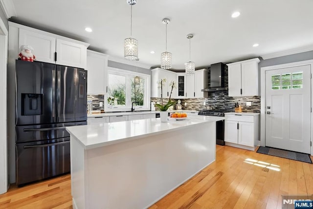 kitchen featuring black refrigerator with ice dispenser, stainless steel gas stove, crown molding, wall chimney range hood, and white cabinets