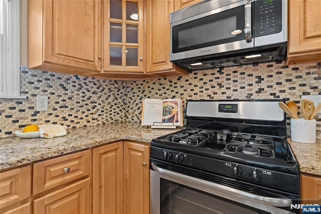 kitchen featuring light stone counters, appliances with stainless steel finishes, glass insert cabinets, and tasteful backsplash