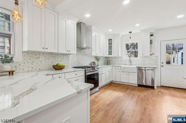 kitchen with pendant lighting, a sink, white cabinetry, appliances with stainless steel finishes, and wall chimney range hood