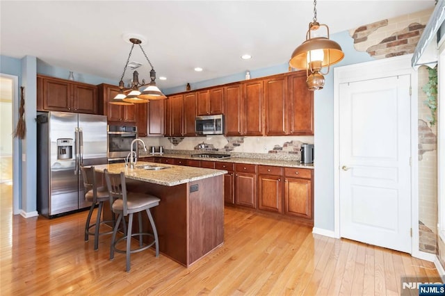 kitchen with a kitchen island with sink, a sink, hanging light fixtures, stainless steel appliances, and light wood-type flooring