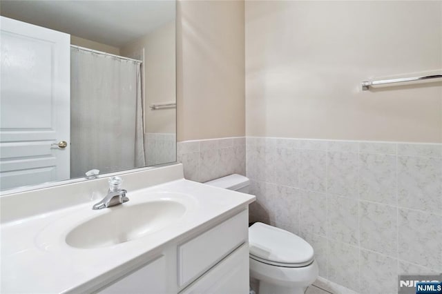 bathroom featuring toilet, a shower with shower curtain, vanity, wainscoting, and tile walls