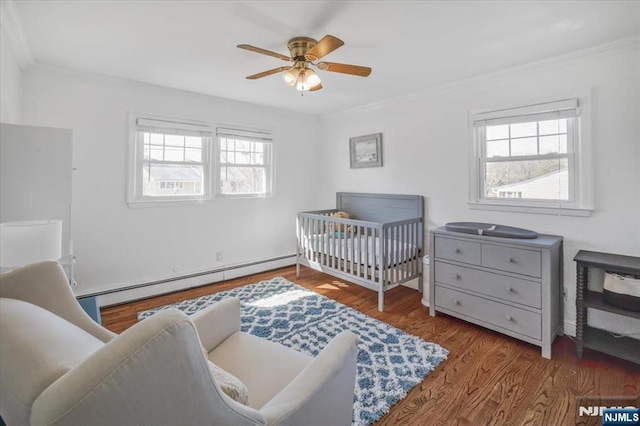 bedroom featuring a crib, a ceiling fan, wood finished floors, crown molding, and baseboard heating