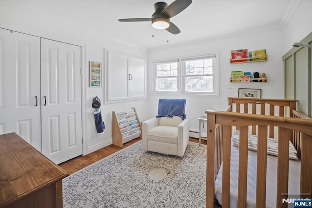 bedroom featuring a crib, wood finished floors, crown molding, a baseboard radiator, and ceiling fan