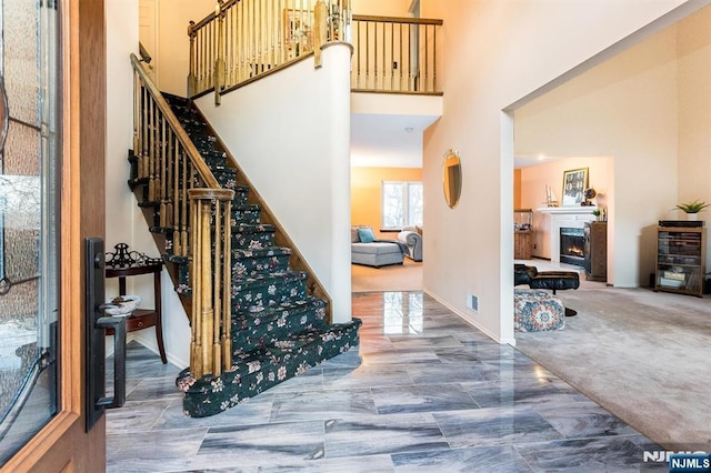 carpeted foyer with stairway, a high ceiling, baseboards, and a warm lit fireplace