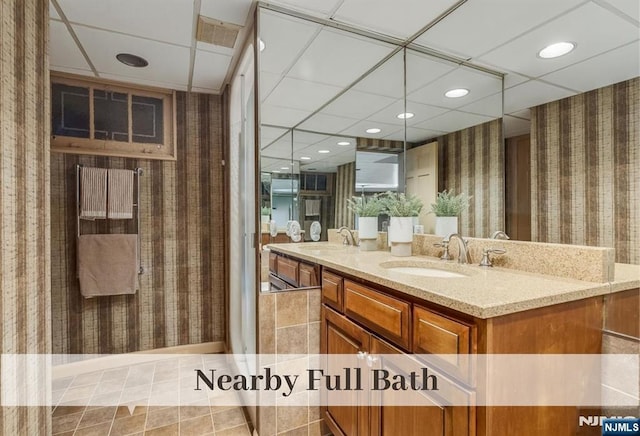 bathroom with wallpapered walls, double vanity, a paneled ceiling, and a sink