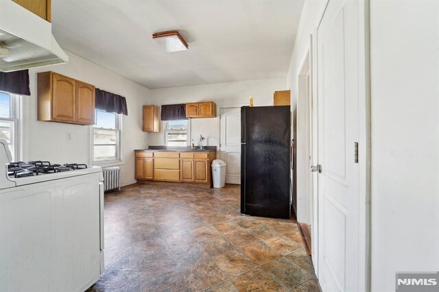 kitchen with radiator heating unit, freestanding refrigerator, white gas range oven, under cabinet range hood, and dark countertops