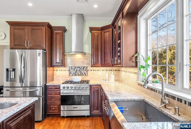 kitchen with light stone countertops, a sink, stainless steel appliances, crown molding, and wall chimney range hood