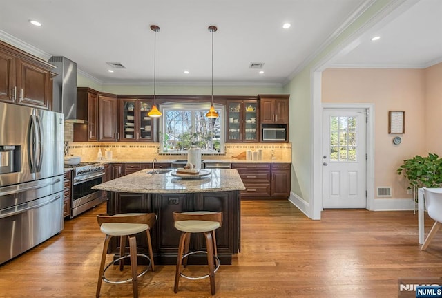 kitchen with wall chimney range hood, light stone countertops, an island with sink, wood finished floors, and stainless steel appliances