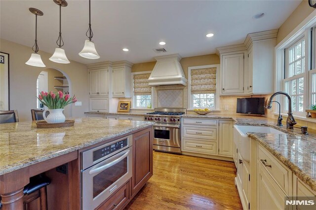 kitchen featuring light wood-type flooring, custom range hood, light stone counters, a wealth of natural light, and appliances with stainless steel finishes