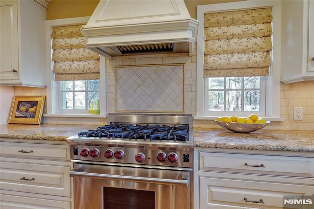 kitchen featuring tasteful backsplash, light stone countertops, custom exhaust hood, high end range, and white cabinetry