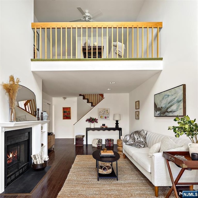 living area with a ceiling fan, a warm lit fireplace, wood finished floors, a high ceiling, and stairs