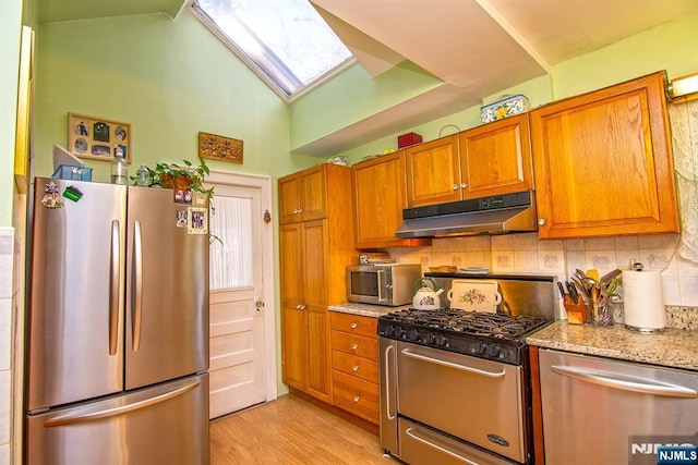 kitchen with brown cabinetry, vaulted ceiling with skylight, stainless steel appliances, decorative backsplash, and under cabinet range hood