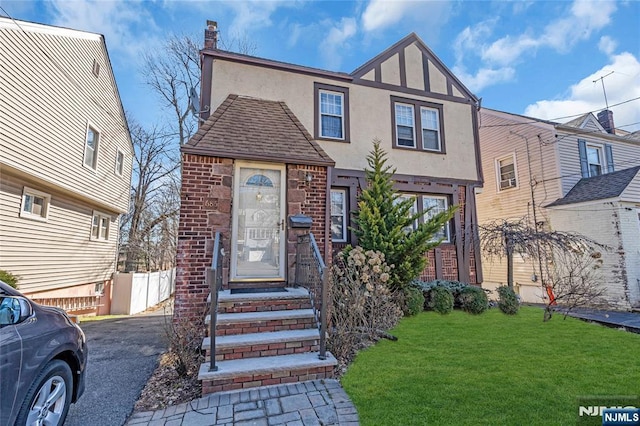 english style home featuring brick siding, a shingled roof, fence, a front yard, and stucco siding