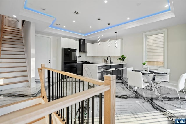 kitchen featuring a tray ceiling, white cabinetry, black appliances, and wall chimney range hood