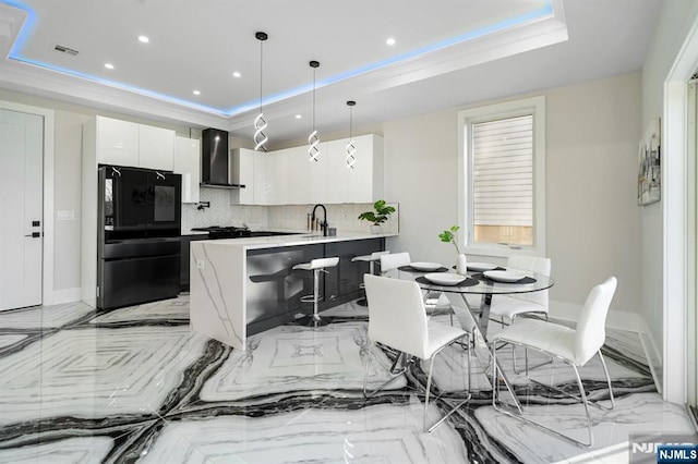 kitchen featuring wall chimney range hood, white cabinets, freestanding refrigerator, marble finish floor, and a raised ceiling