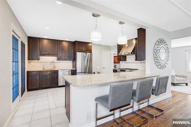 kitchen with dark brown cabinetry, light stone counters, appliances with stainless steel finishes, and wall chimney range hood