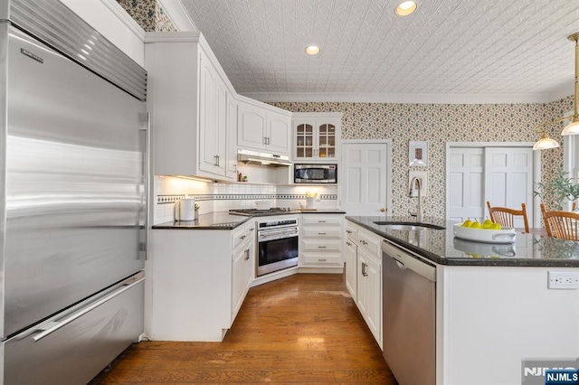 kitchen featuring wood finished floors, wallpapered walls, a sink, under cabinet range hood, and built in appliances