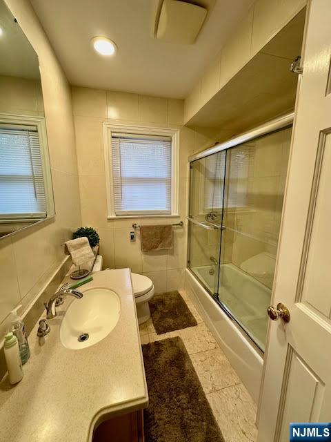 bathroom featuring combined bath / shower with glass door, toilet, vanity, and tile walls