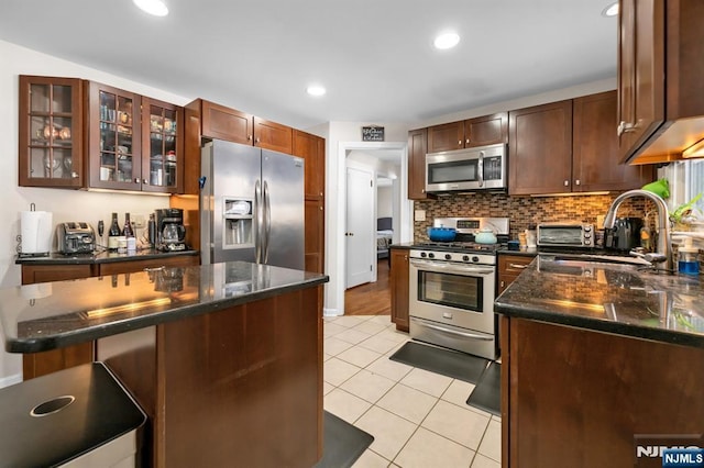 kitchen with glass insert cabinets, light tile patterned floors, decorative backsplash, stainless steel appliances, and a sink