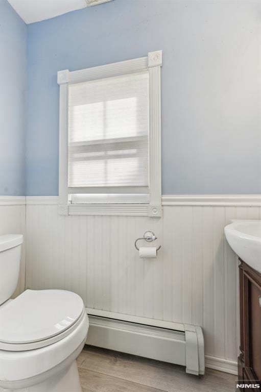 half bath with vanity, wood finished floors, a baseboard radiator, wainscoting, and toilet