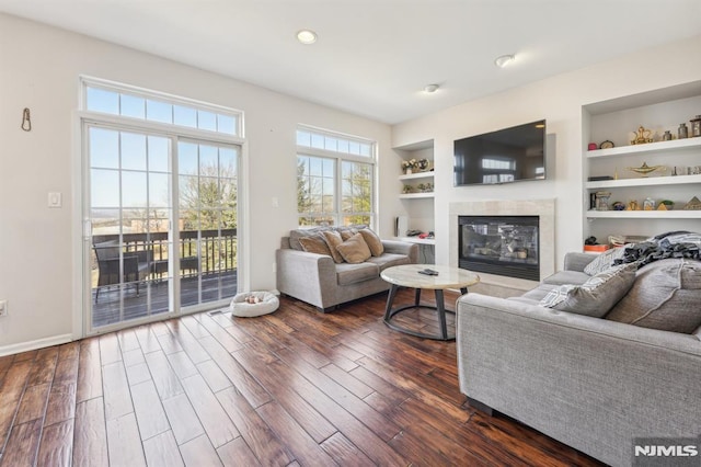 living area featuring dark wood-type flooring, baseboards, built in features, recessed lighting, and a glass covered fireplace