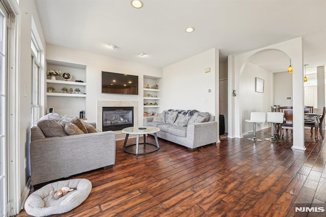 living area featuring built in shelves, recessed lighting, arched walkways, a glass covered fireplace, and dark wood-style flooring