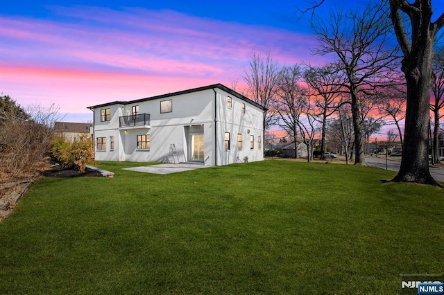 back of property at dusk with a patio area, a balcony, a lawn, and stucco siding