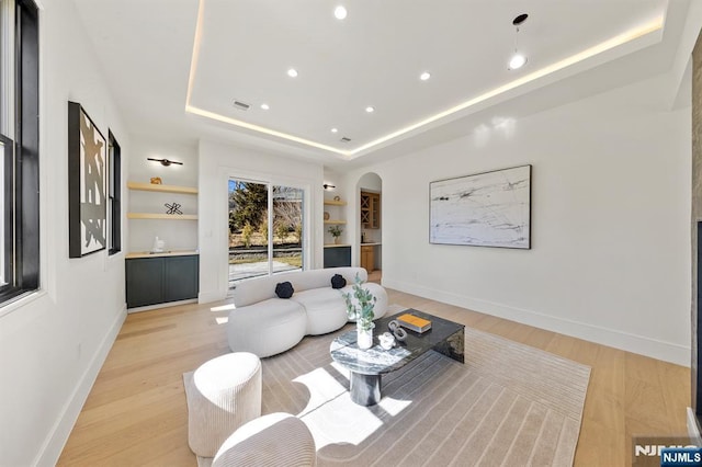 living room with built in features, baseboards, a tray ceiling, arched walkways, and light wood-style floors