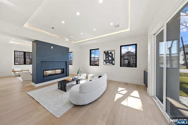 living area with visible vents, a tray ceiling, a multi sided fireplace, light wood-style floors, and baseboards