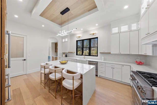 kitchen with light wood-type flooring, a kitchen bar, a tray ceiling, arched walkways, and appliances with stainless steel finishes