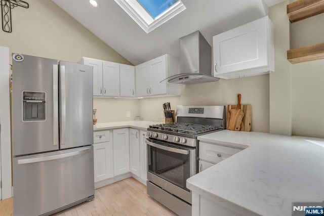 kitchen with vaulted ceiling with skylight, appliances with stainless steel finishes, white cabinetry, wall chimney exhaust hood, and light wood-type flooring