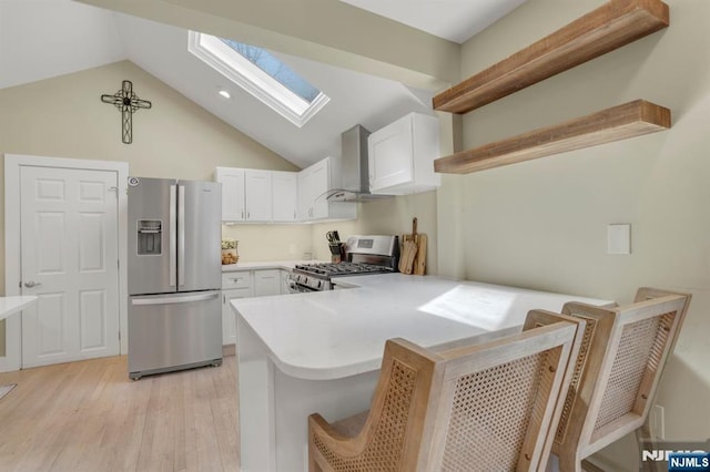 kitchen featuring stainless steel appliances, a peninsula, a skylight, white cabinets, and wall chimney range hood