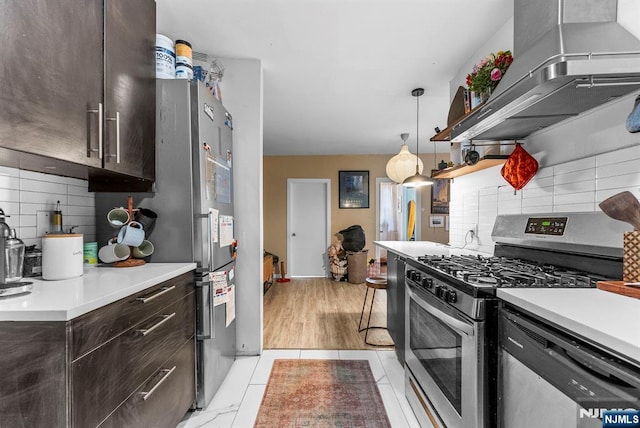 kitchen with stainless steel appliances, decorative backsplash, light countertops, dark brown cabinets, and wall chimney exhaust hood