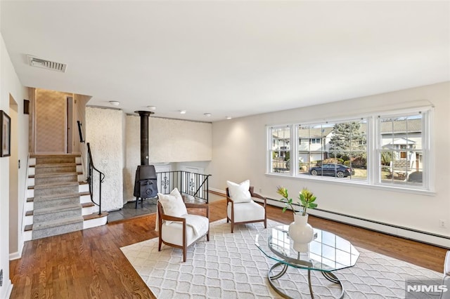 sitting room featuring visible vents, wood finished floors, stairway, a baseboard radiator, and a wood stove