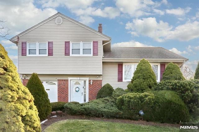 split level home featuring a garage, brick siding, a chimney, and a shingled roof