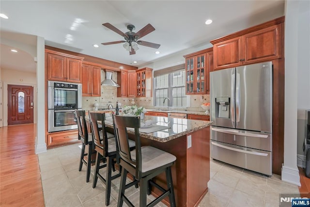 kitchen with light stone counters, glass insert cabinets, appliances with stainless steel finishes, wall chimney exhaust hood, and backsplash