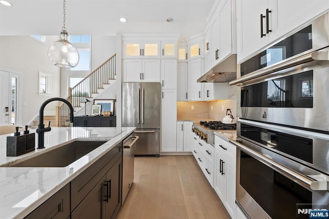 kitchen with stainless steel appliances, under cabinet range hood, a sink, light stone counters, and hanging light fixtures