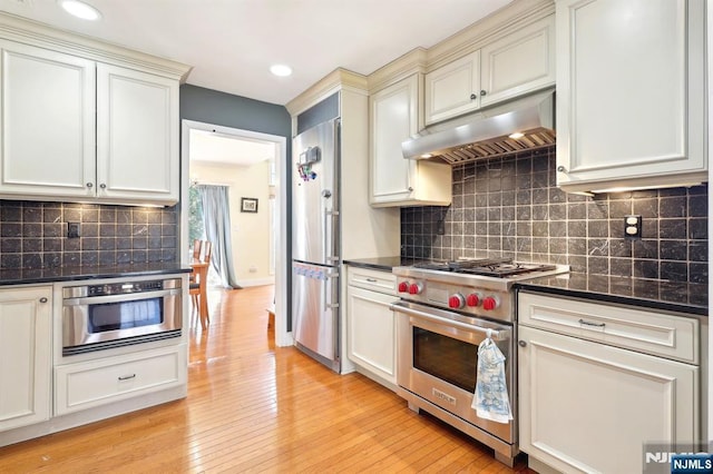 kitchen featuring high end appliances, tasteful backsplash, light wood-style flooring, under cabinet range hood, and dark stone counters