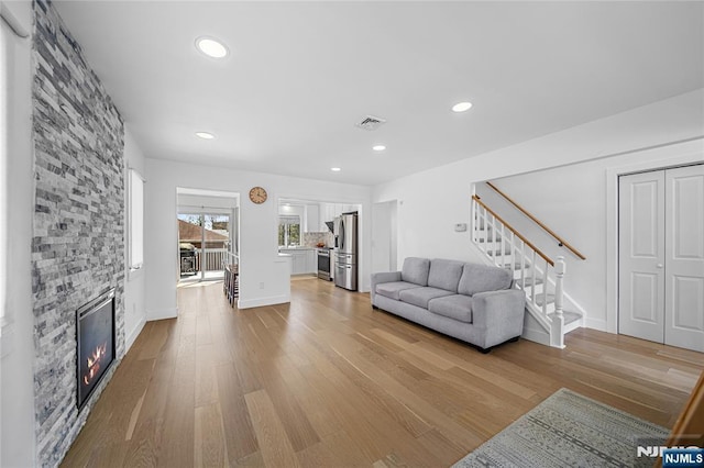living room featuring a stone fireplace, stairway, baseboards, recessed lighting, and light wood-style flooring