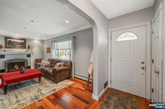 foyer with crown molding, a brick fireplace, arched walkways, wood finished floors, and a baseboard heating unit