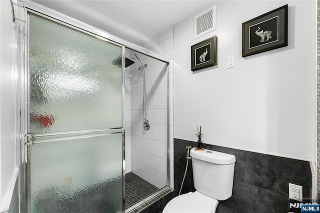 bathroom with tile walls, toilet, a shower stall, a wainscoted wall, and visible vents