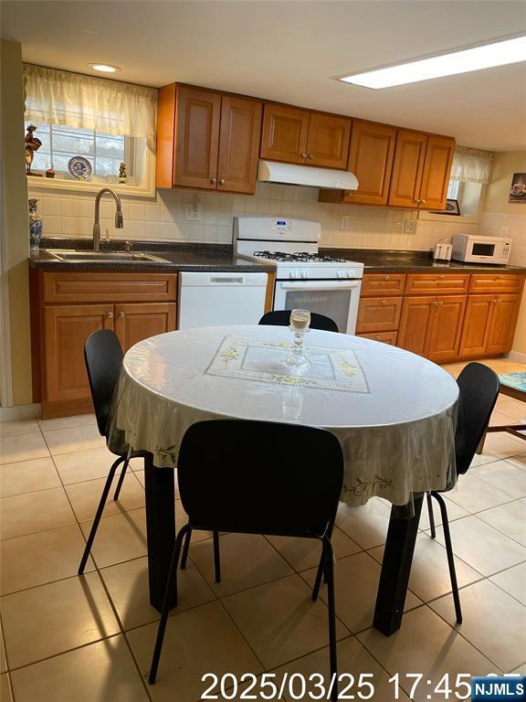 kitchen featuring a sink, tasteful backsplash, brown cabinets, under cabinet range hood, and white appliances