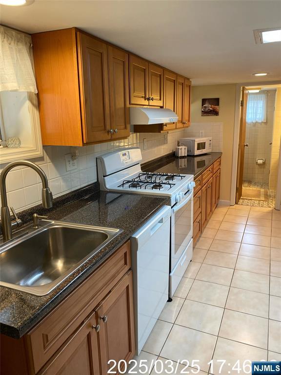 kitchen with light tile patterned flooring, white appliances, brown cabinets, under cabinet range hood, and a sink