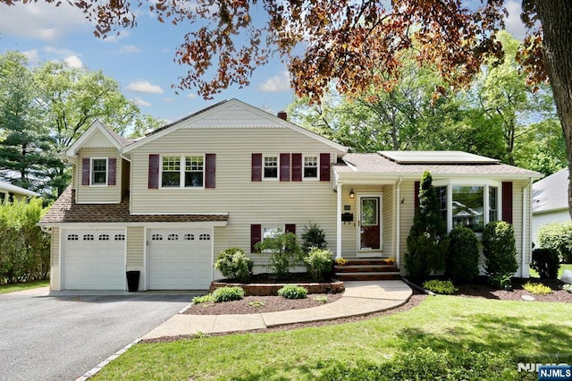 tri-level home with asphalt driveway, solar panels, a garage, a front lawn, and roof with shingles