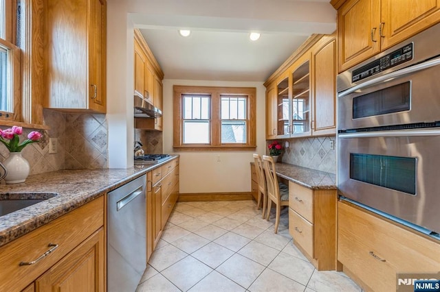 kitchen featuring light tile patterned floors, glass insert cabinets, appliances with stainless steel finishes, baseboards, and under cabinet range hood