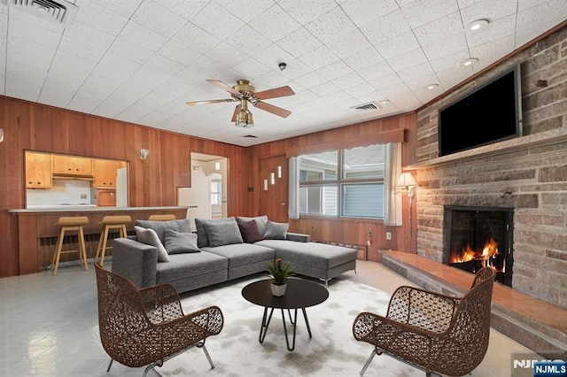 living room featuring wooden walls, ceiling fan, a stone fireplace, and visible vents