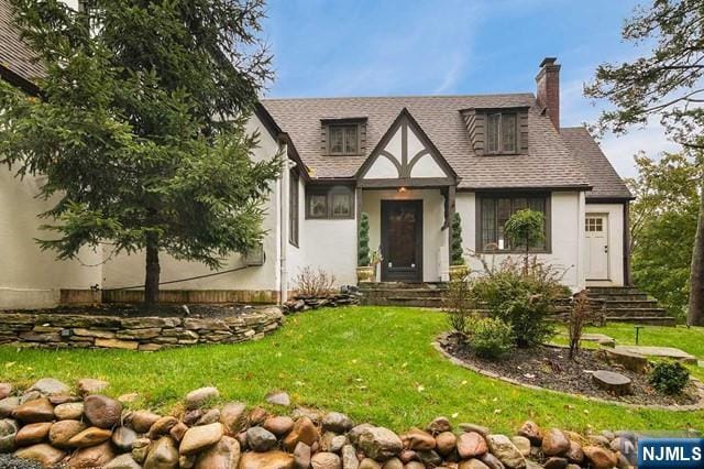 english style home with a chimney, a shingled roof, stucco siding, and a front lawn