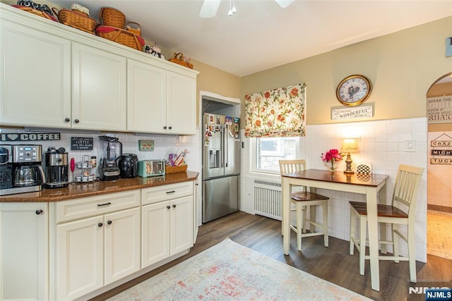 kitchen featuring dark stone countertops, radiator, wood finished floors, ceiling fan, and stainless steel refrigerator with ice dispenser