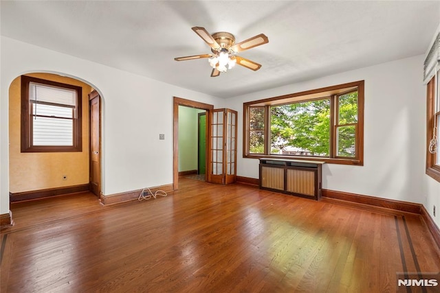 empty room with radiator, wood finished floors, baseboards, arched walkways, and a ceiling fan