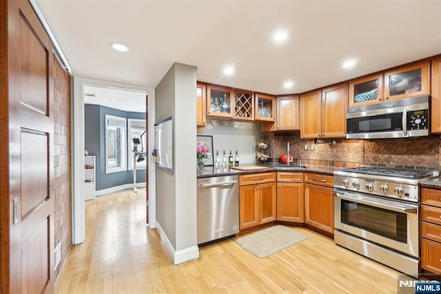kitchen featuring decorative backsplash, glass insert cabinets, light wood-style floors, brown cabinets, and stainless steel appliances